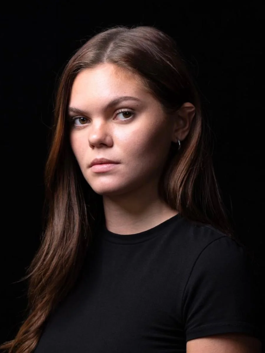 Jeune femme 'Sasha Gravat' aux cheveux longs et bruns, portant un t-shirt noir, regardant sérieusement, fond noir.