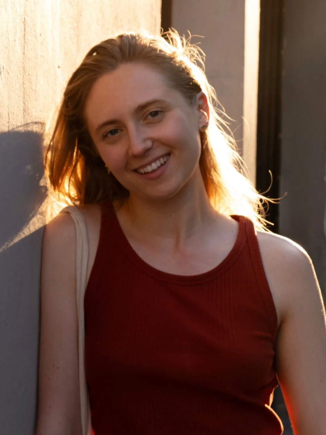 Une jeune femme 'Manon Remadi' souriante avec des cheveux bruns clairs, portant un haut sans manches rouge, debout près d'un mur au coucher du soleil.