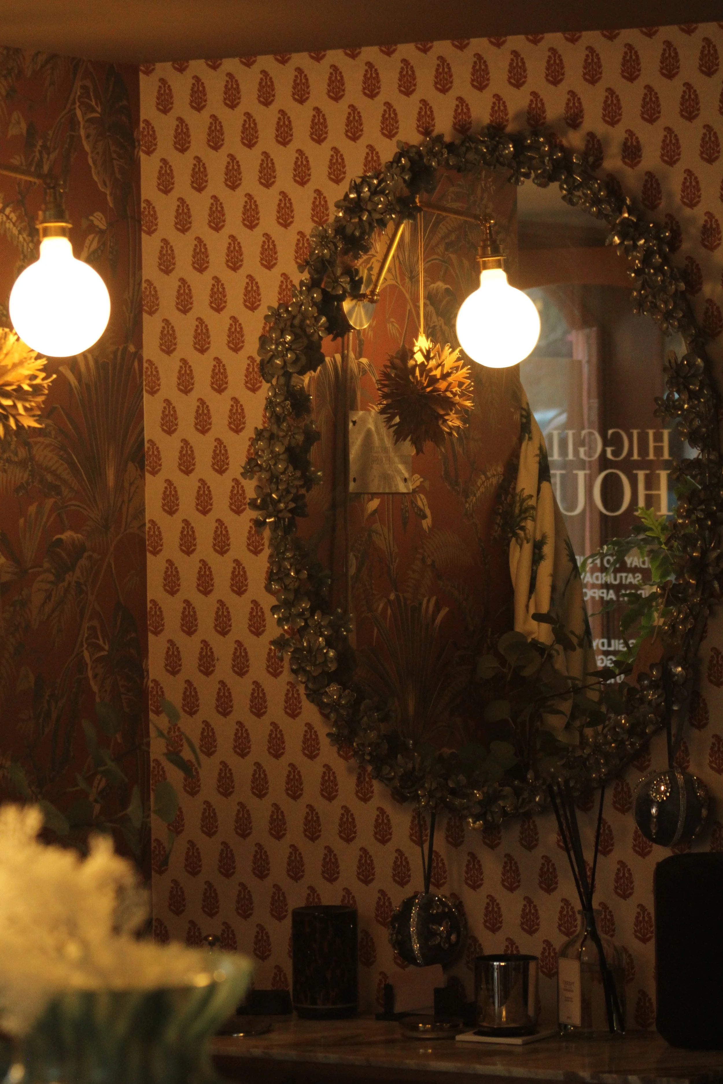 A decorated interior wall with a large oval mirror trimmed with flowers, two lit round bulbs, patterned wallpaper, and various small decorative objects on a table.