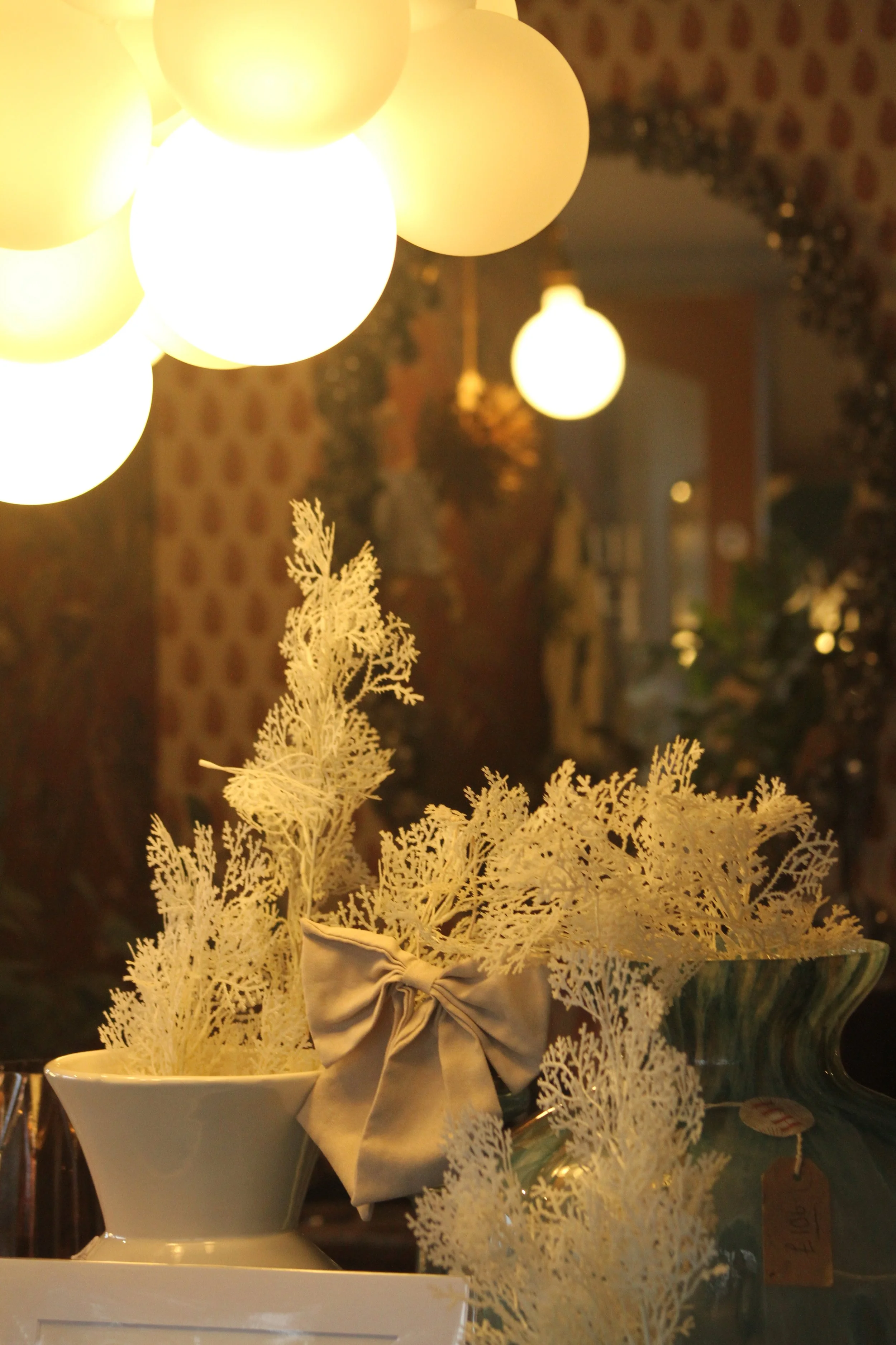 Decorative indoor scene featuring a white vase with faux white foliage and a beige satin bow, illuminated by warm yellow hanging globe lights. In the background, there is a mirror, patterned wallpaper, and various plants and ornaments.