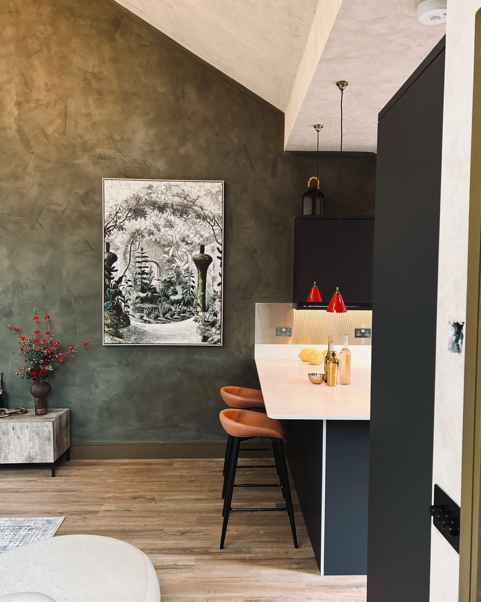 Interior of a modern kitchen area with green textured wall, wall art, wooden flooring, a small side table with a flower vase, and a bar counter with two orange stools and red pendant lights.