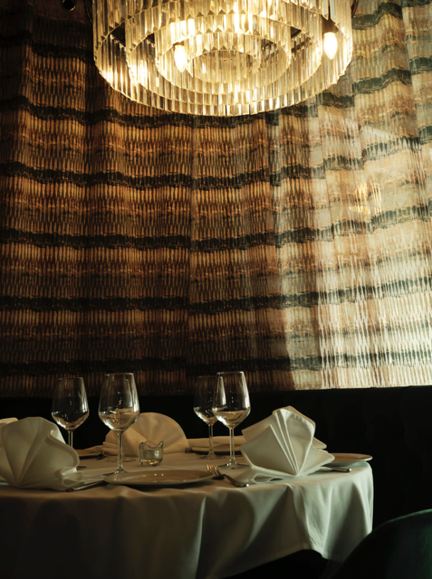 Elegant restaurant table set with three wine glasses, folded white napkins, and a white tablecloth, illuminated by a modern chandelier hanging from a textured, patterned wall.
