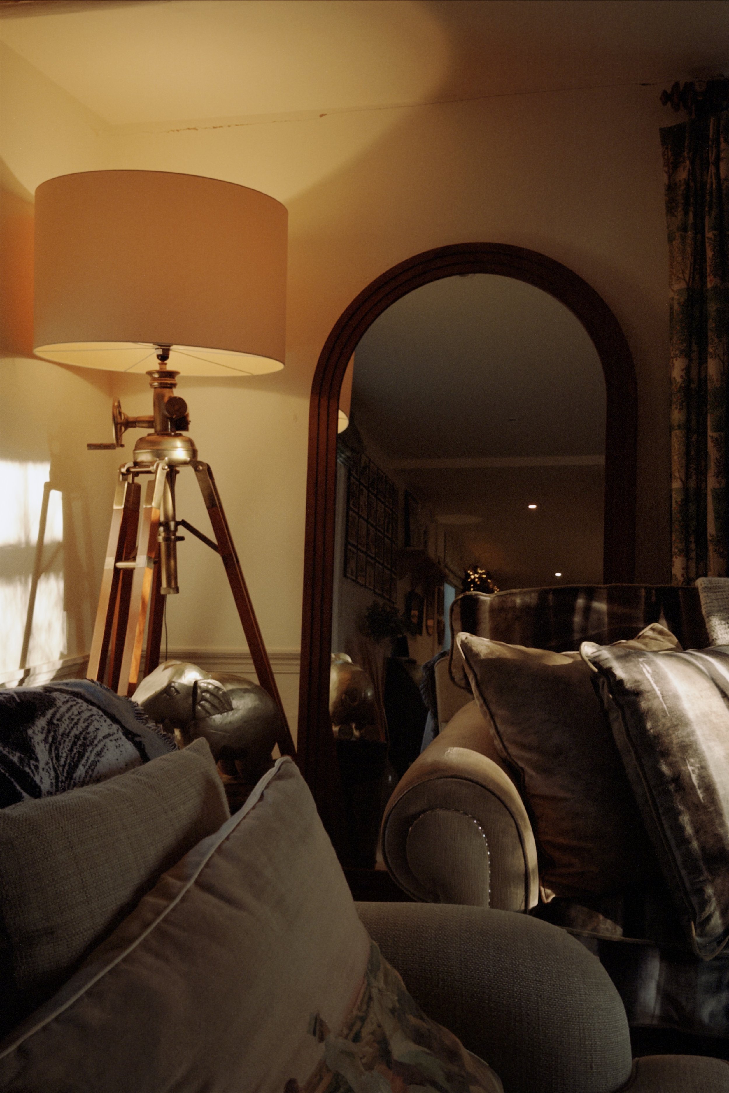 A cozy living room with a wooden arch doorway, a beige sofa with pillows, a large floor lamp with a tripod base, and a mirror reflecting part of the room.