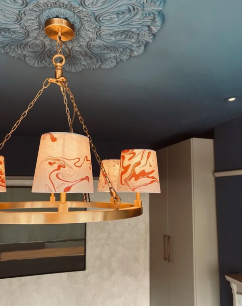 A modern chandelier with three lampshades suspended from a gold chain, hanging from a ceiling medallion with intricate design, in a contemporary room.