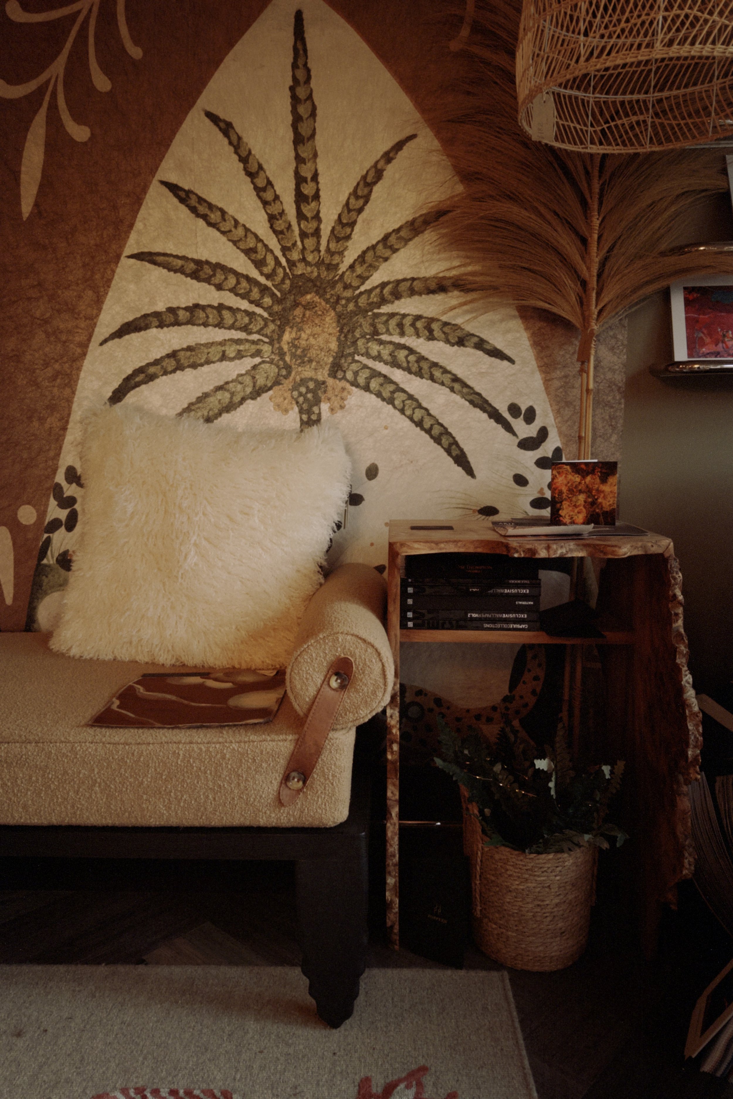 A cozy living room corner with a beige upholstered armchair, a fluffy cream-colored pillow, a wooden side table with books, a framed picture, and a decorative plant in a woven basket, all set against a wall with a palm leaf mural.