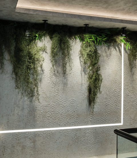 Interior wall with a textured surface, decorated with hanging green plants and black wall-mounted lights. A white LED strip light runs along the wall's corners.