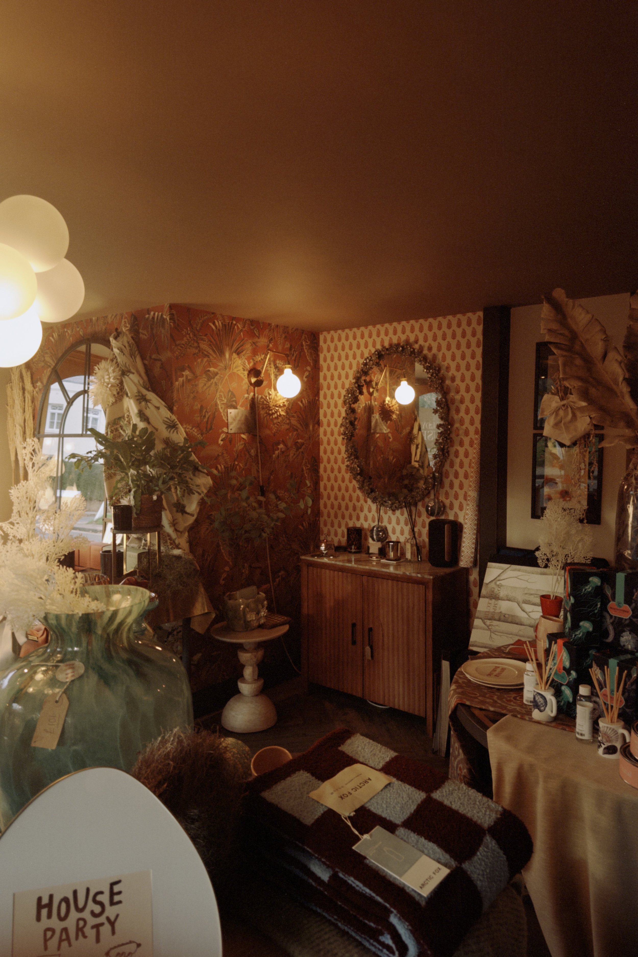 Cozy, warmly lit interior of a room decorated with vintage and eclectic decor, including a large mirror with a floral frame, patterned wallpaper, and various decorative objects on tables and shelves.