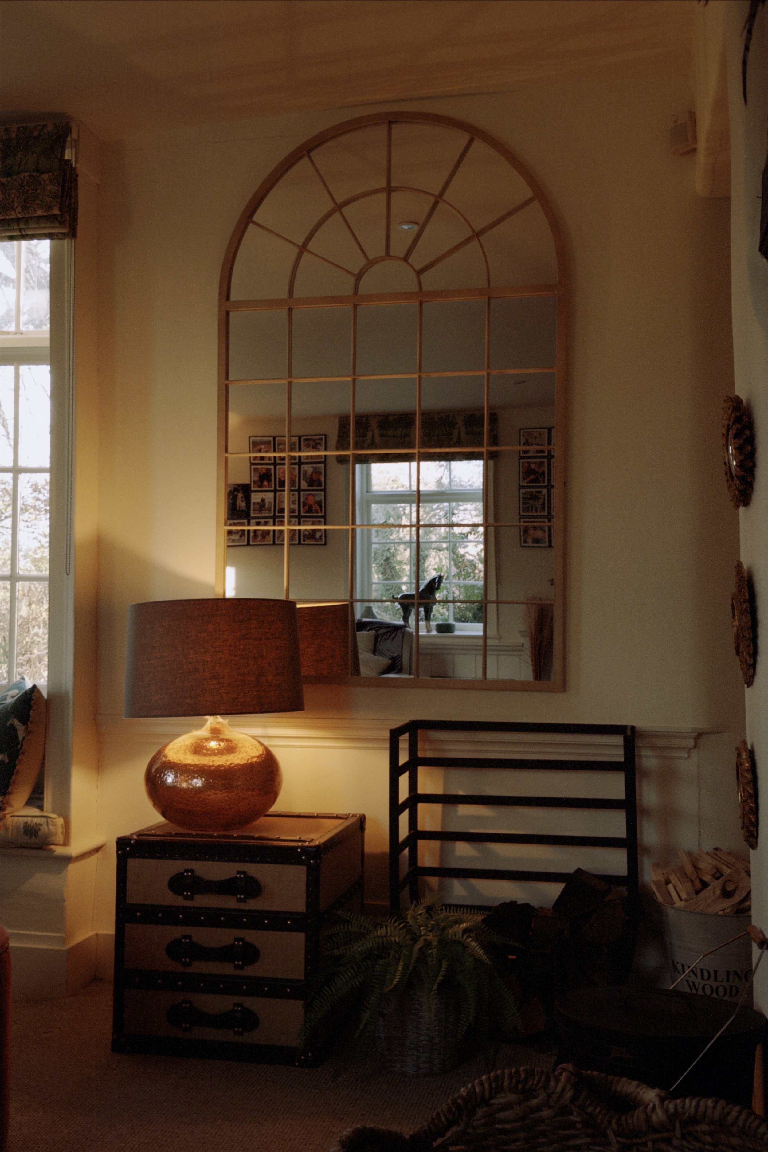 Living room with a large decorative mirror, a table lamp on a wooden and metal chest, a window with natural light, and various decorations.