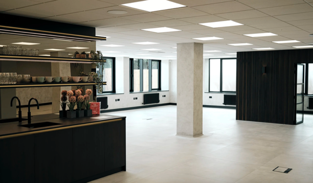 An interior space with large windows, black furniture, open shelving, and a kitchen island with flowers and dishes.