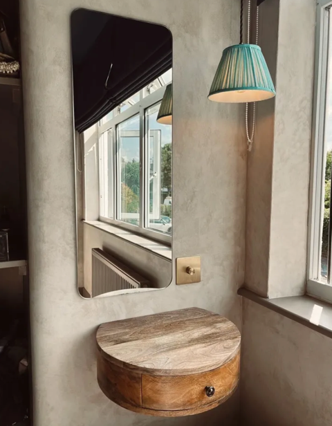 Close-up of a rounded wooden floating shelf beneath a tall mirror, with a window and two teal-colored pendant lights in the background.