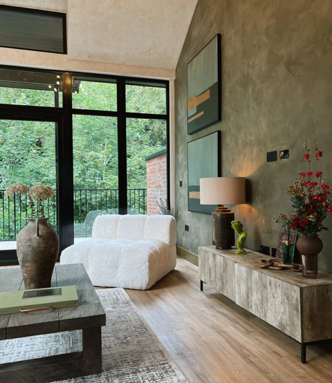 Modern living room with large windows, a white sofa, abstract wall art, and a textured sideboard with decorative vases and flowers.