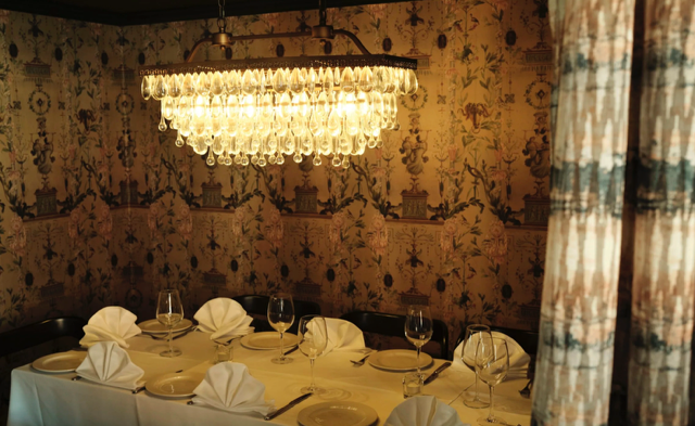 Elegant dining room with a long table set with plates, wine glasses, and folded napkins, illuminated by a large chandelier, with patterned wallpaper and curtains.