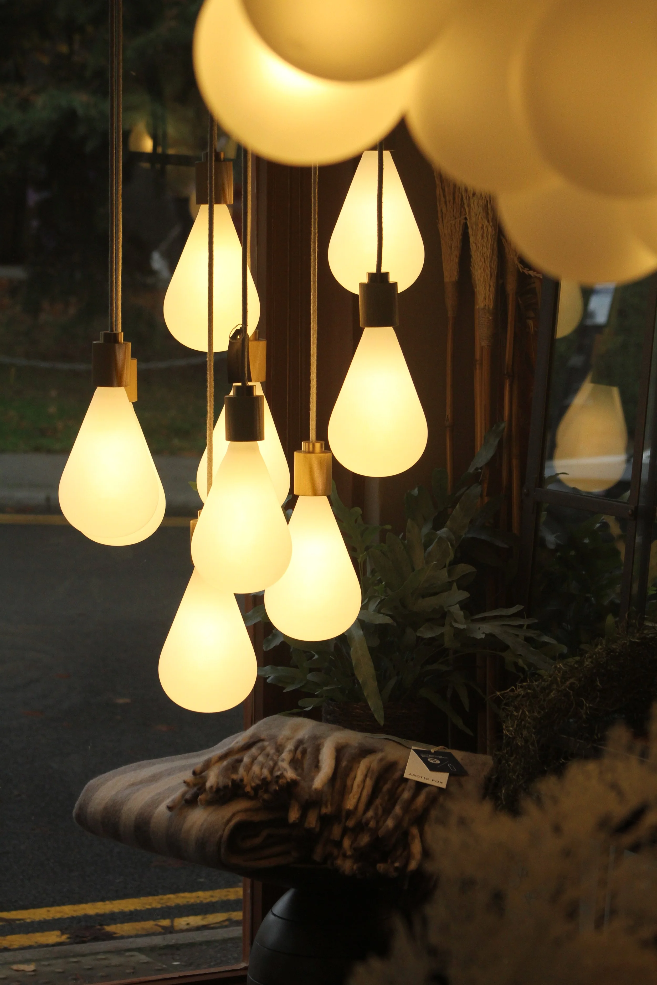 Ceiling lights shaped like teardrops hanging in a store window, with some indoor plants and a folded blanket on a small table nearby.