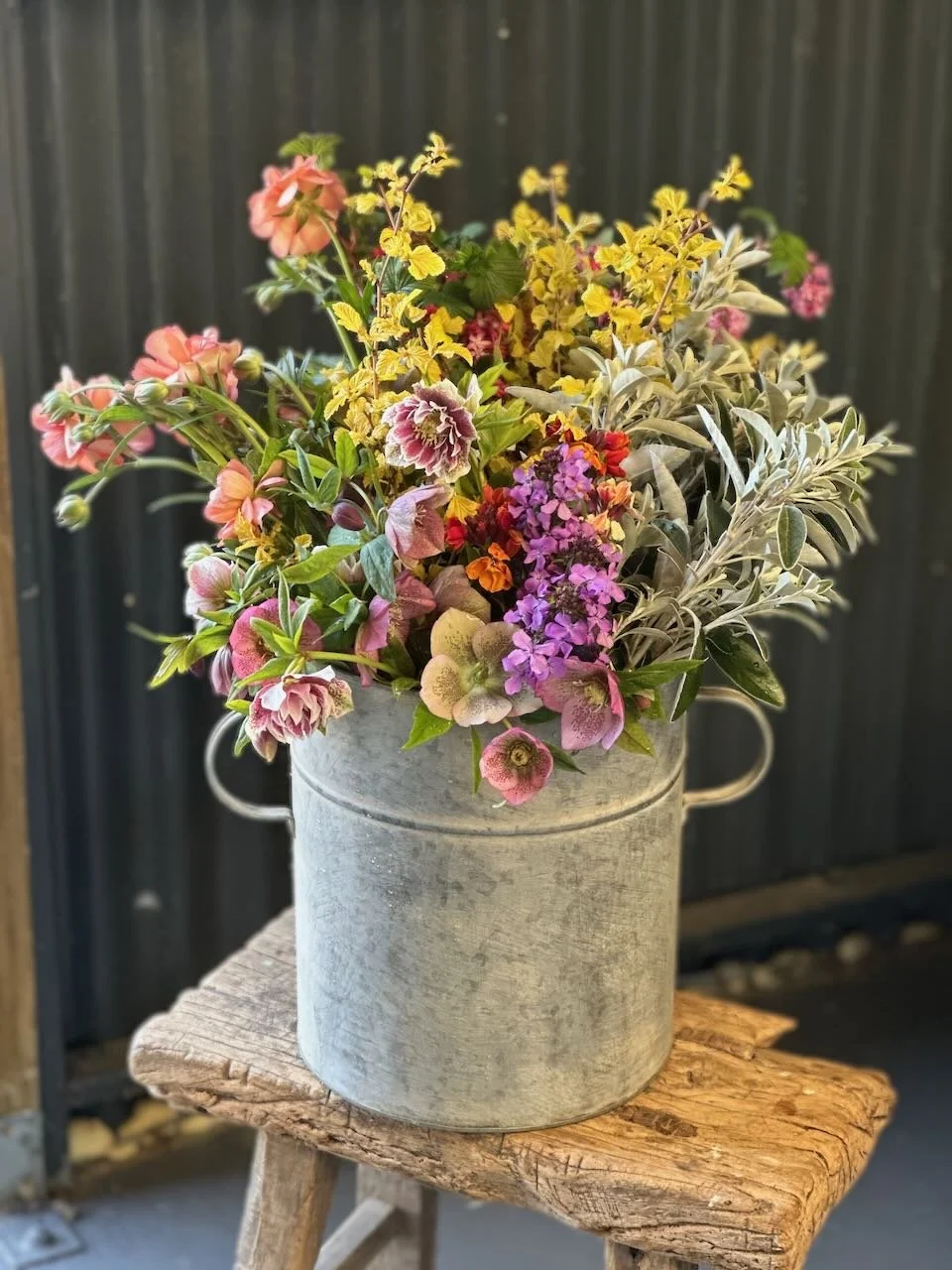 A rustic gray metal bucket filled with colorful flowers, including pink, yellow, purple, and white blossoms, sitting on a weathered wooden stool outdoors with a dark vertical fence in the background.