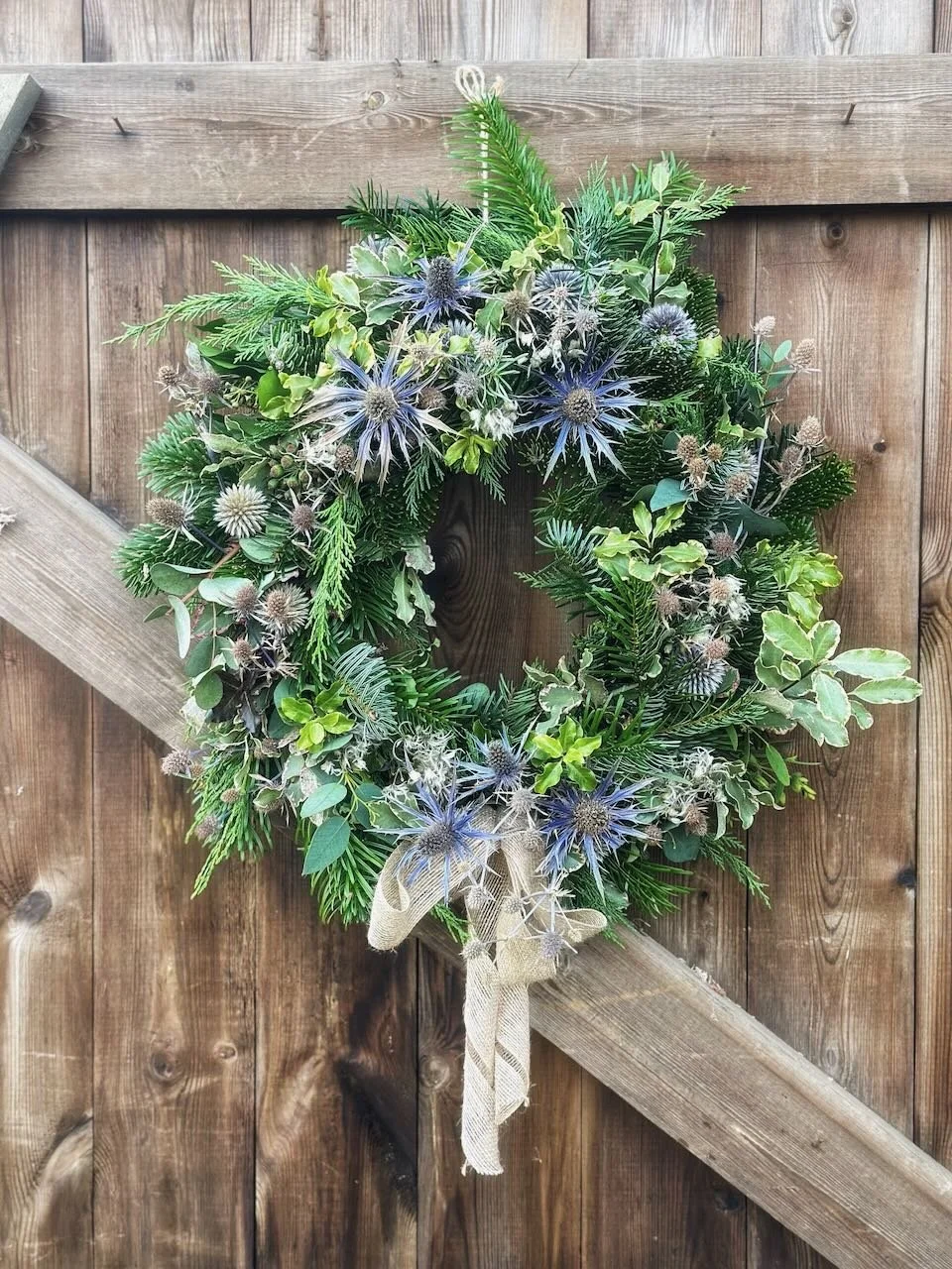 A decorative wreath made of various green leaves and blue flowers, hanging on a wooden fence with a burlap ribbon bow.