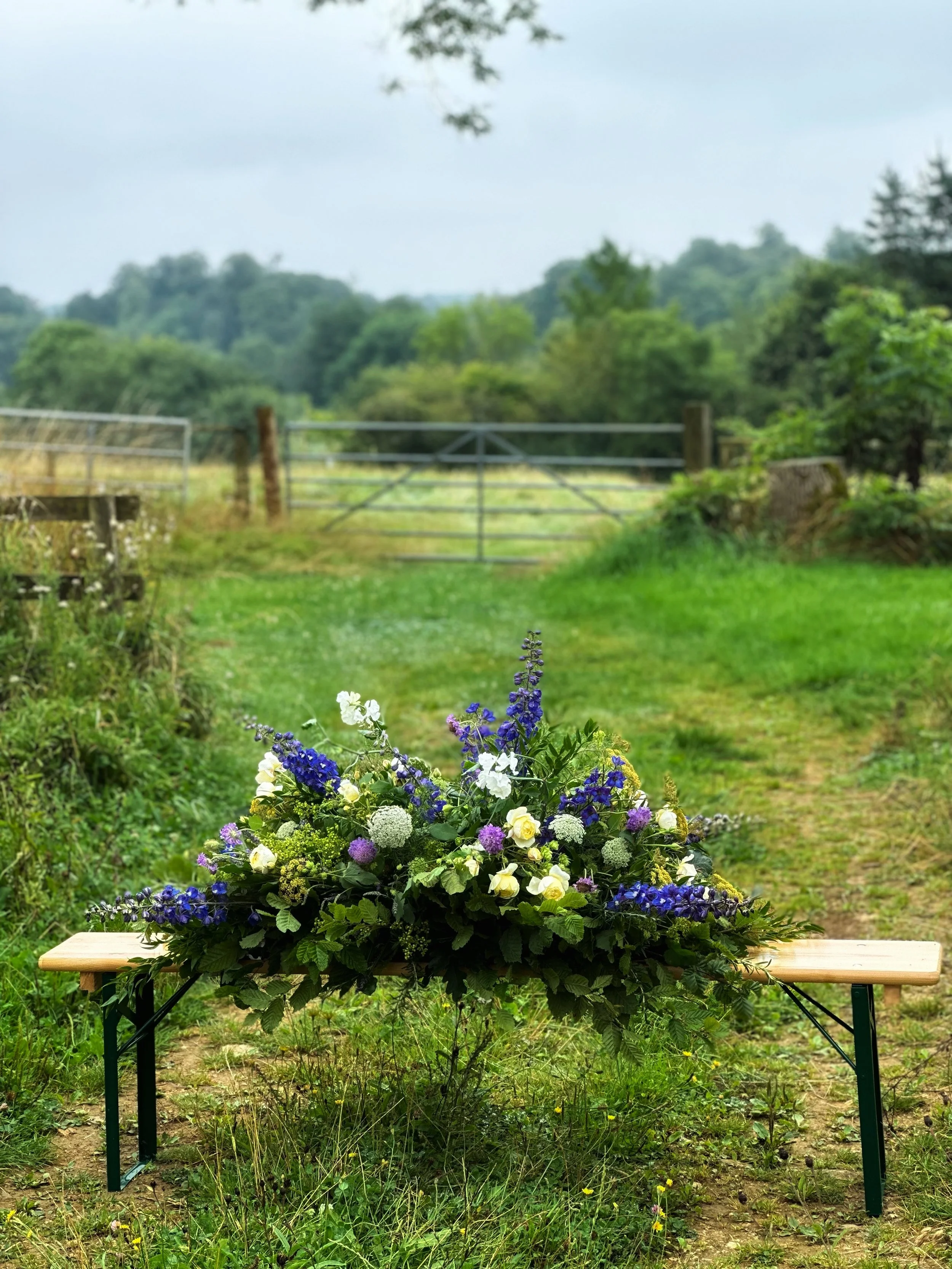 A colorful flower arrangement placed on a wooden bench outdoors in a rural setting, with a green grassy field and trees in the background.