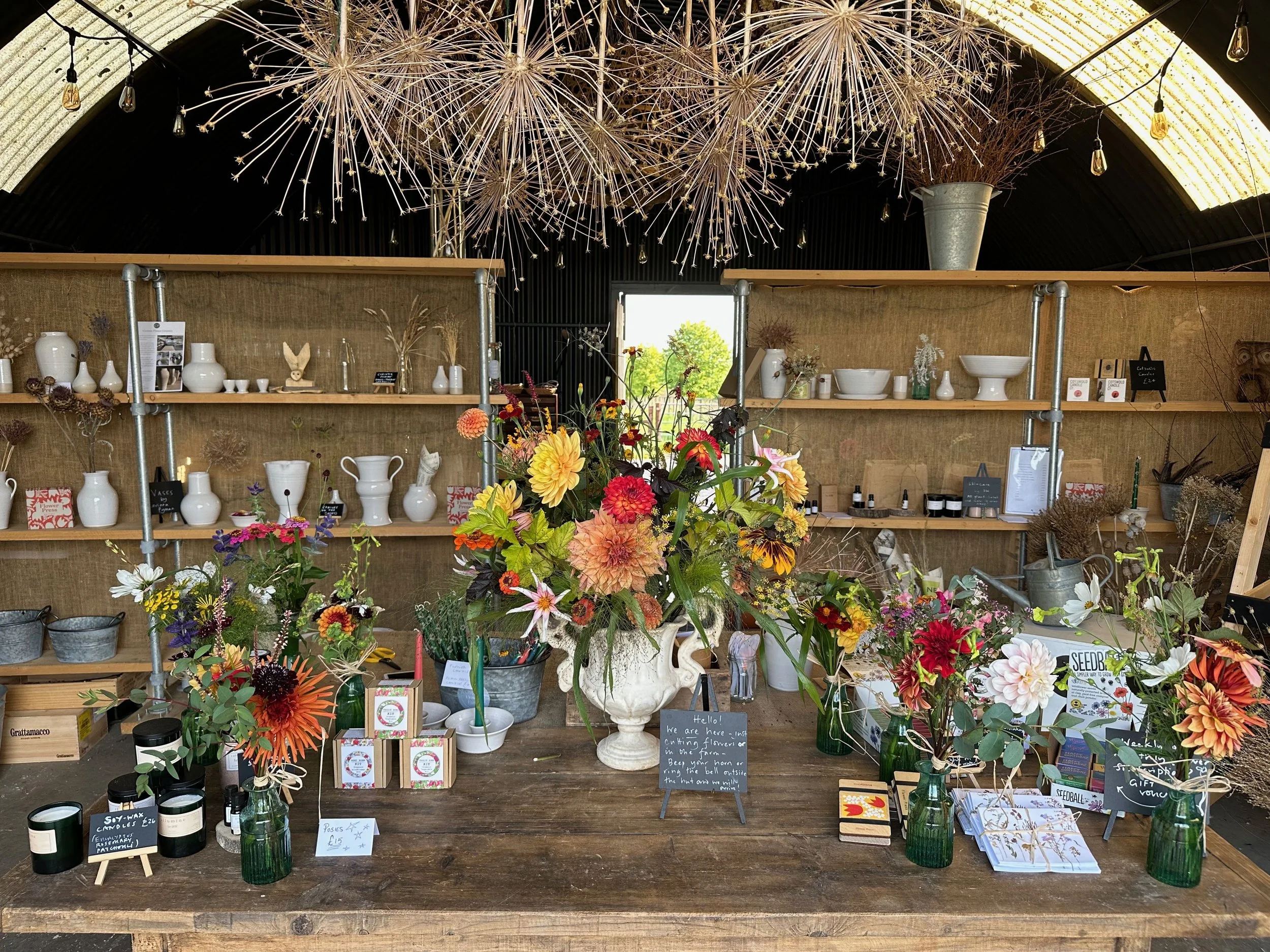 Inside a flower shop with a large central floral arrangement of colorful flowers in a white vase, surrounded by smaller flower arrangements, flower care products, and décor items on a wooden table. Shelves in the background hold white vases and decor