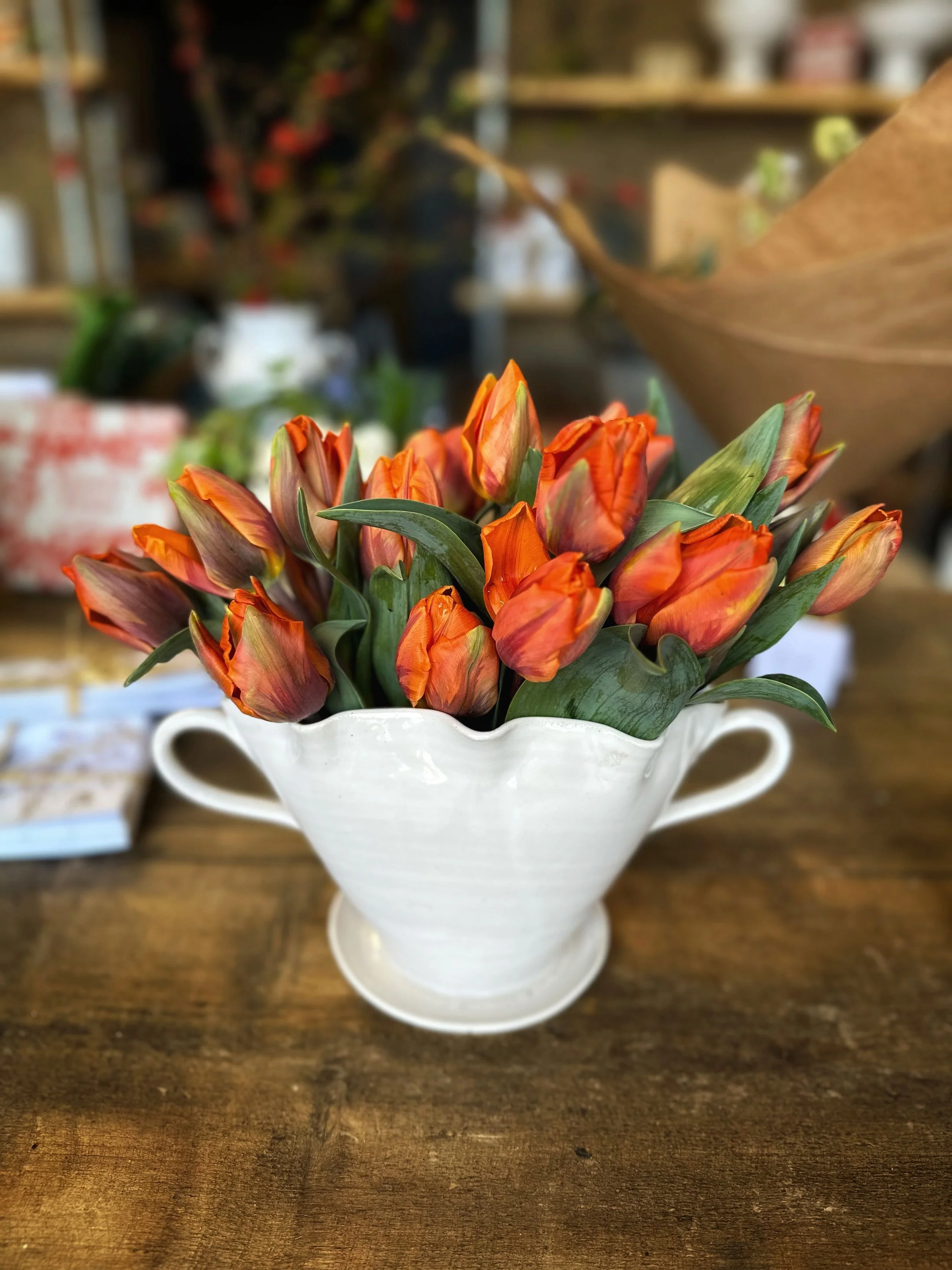 A white ceramic vase filled with orange tulips on a wooden surface.