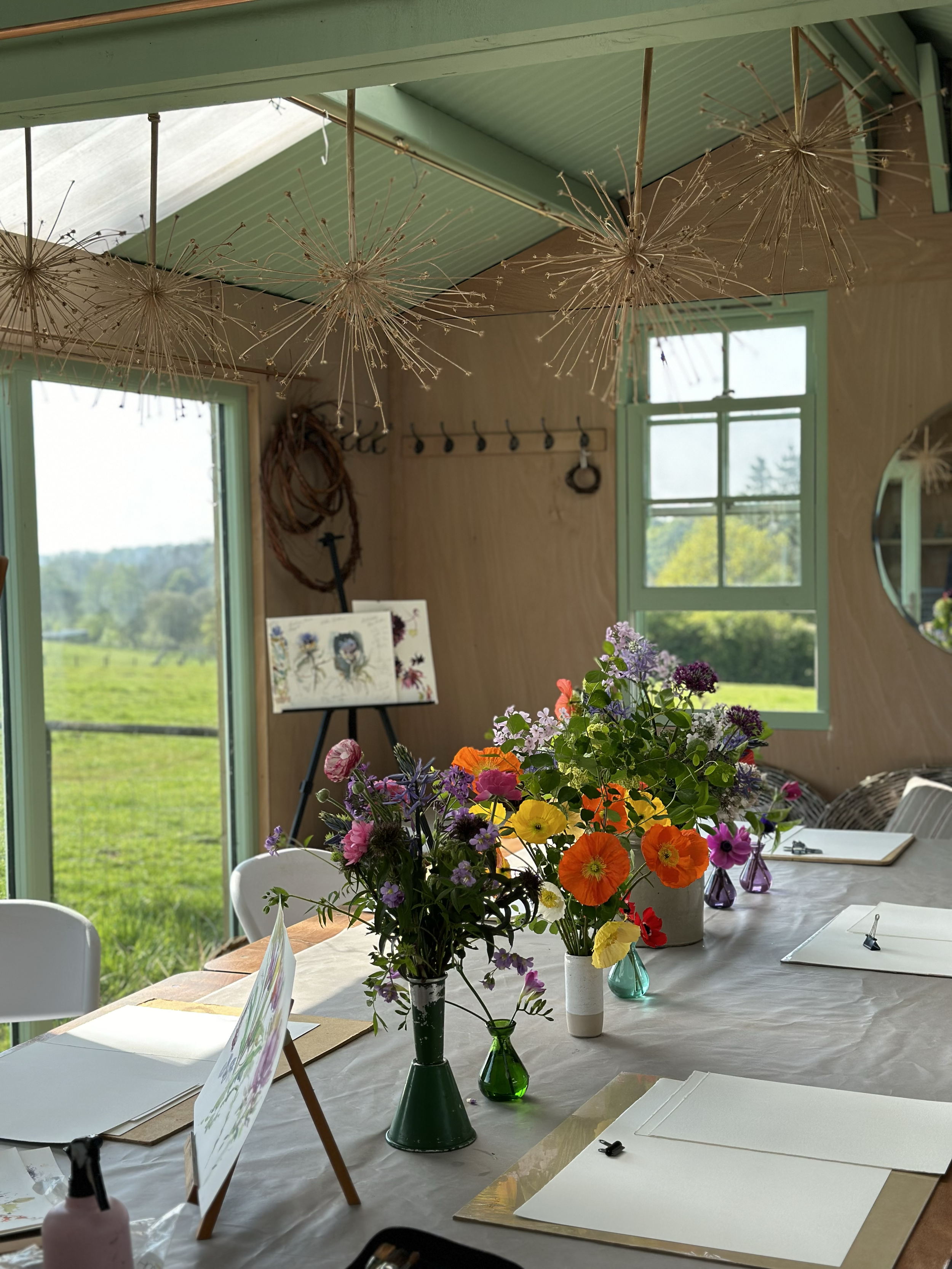 A cozy interior with large windows showing a green outdoor landscape, decorated with hanging dandelion-inspired ornaments, featuring a table with colorful flower arrangements in vases, art supplies, and notebooks.