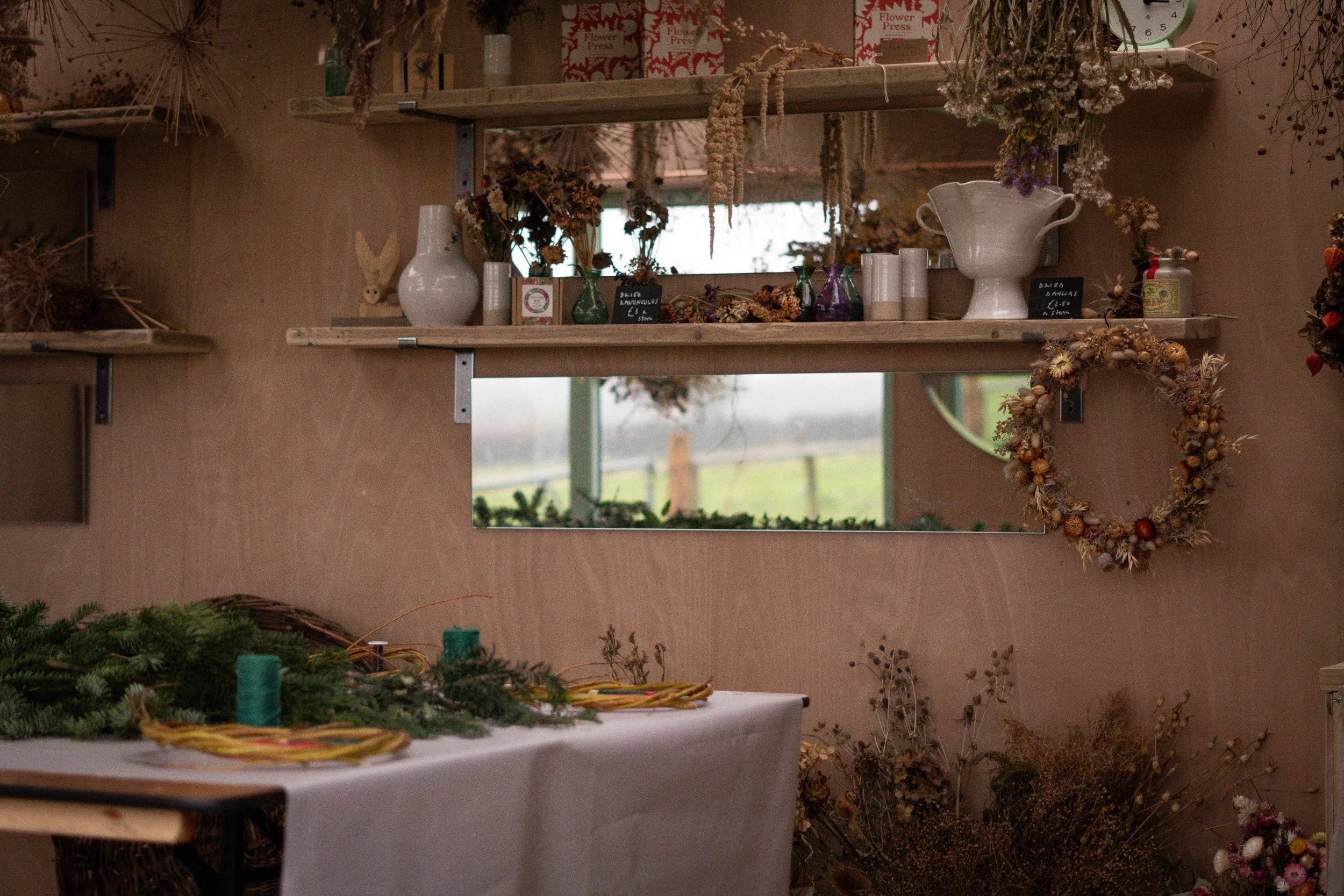 A cozy workshop with shelves filled with dried flowers, vases, and craft supplies. A wreath hangs on the wall, and a table in the foreground holds evergreen branches and candles, suggesting preparations for a holiday or seasonal event.