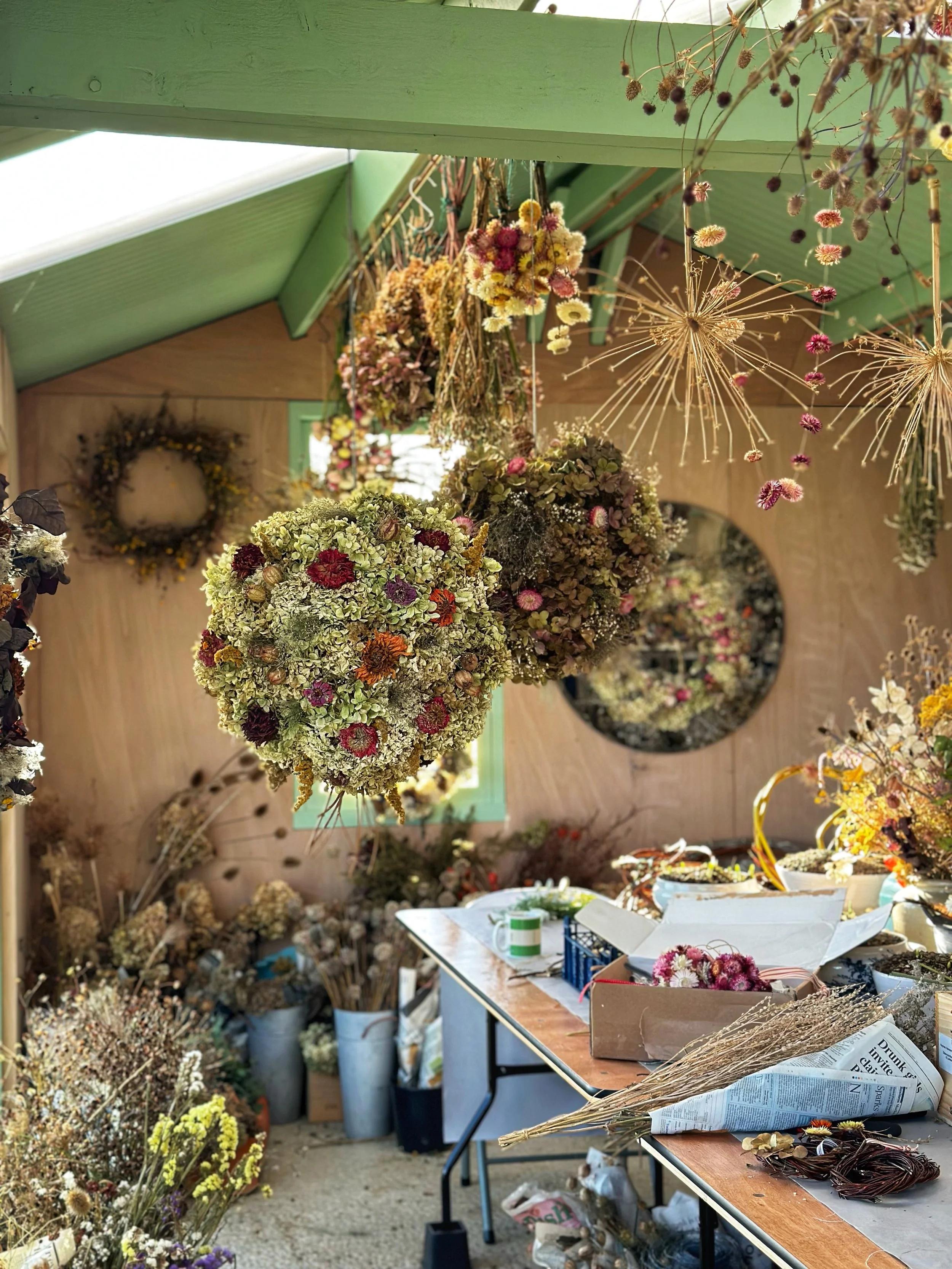 A room with dried flowers hanging from the ceiling, a table with floristry supplies, and a mirror on the back wall. The room has a green ceiling and a window in the background.