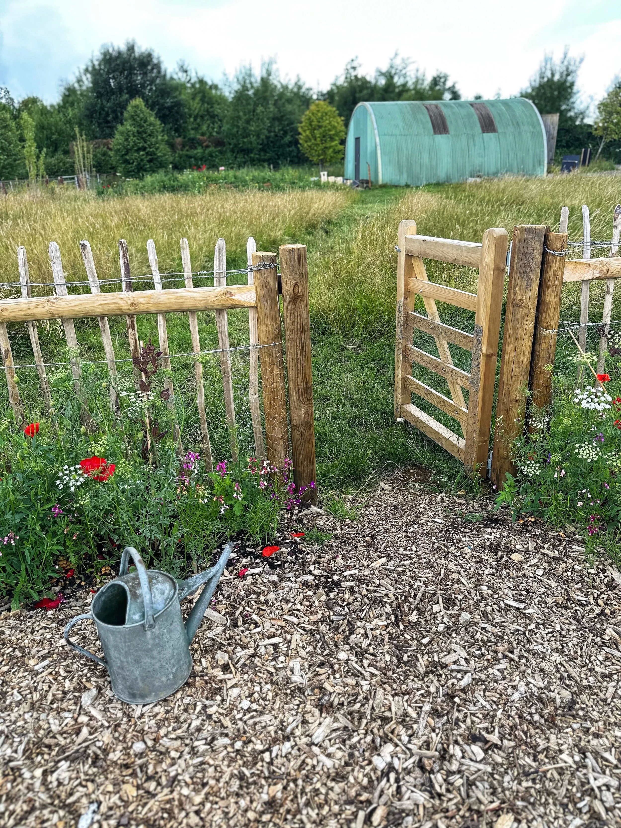 Garden with wooden fence and gate, colorful flowers, watering can, and a greenhouse in the background.