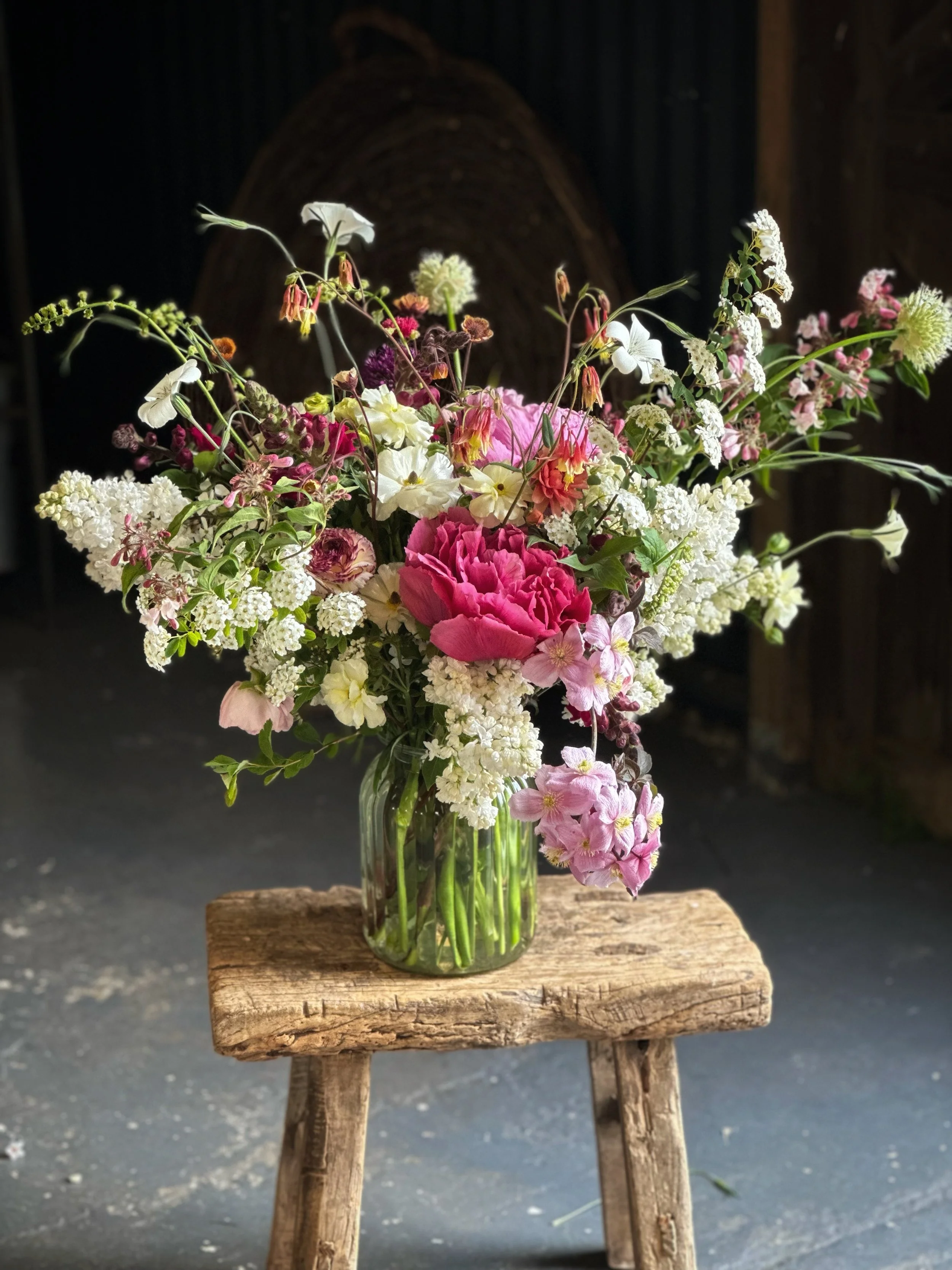 A glass vase filled with a colorful bouquet of flowers, including pink, white, and green blooms, placed on a rustic wooden stool inside a barn or similar structure.