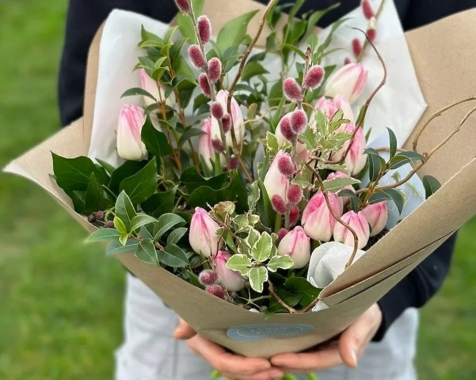 A person holding a bouquet of pink and white tulips, greenery, and fuzzy pink catkins wrapped in brown paper outside on green grass.
