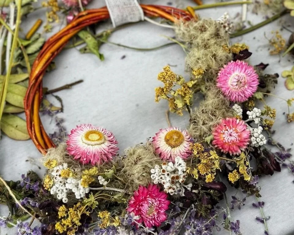 A floral wreath made of pink and white flowers, dried yellow flowers, moss, and branches, resting on a white surface.