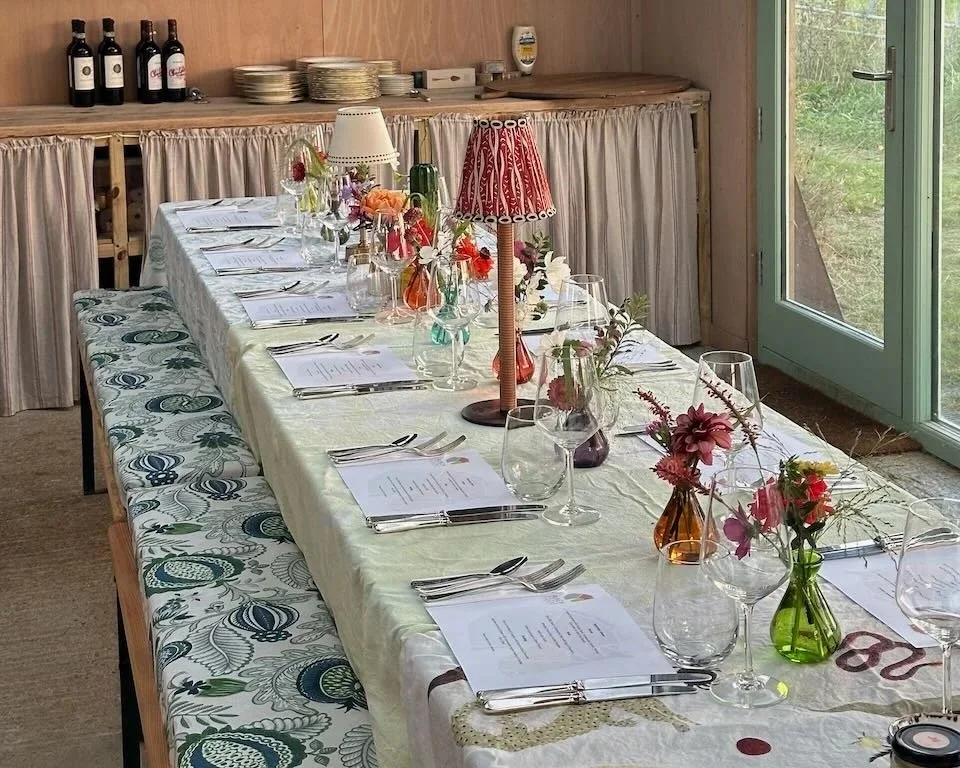 A decorated dining table set for a meal with flowers in small vases, candle lamps, and place settings with papers, forks, knives, and glasses, in a cozy room with a sliding glass door and bottles on a shelf.