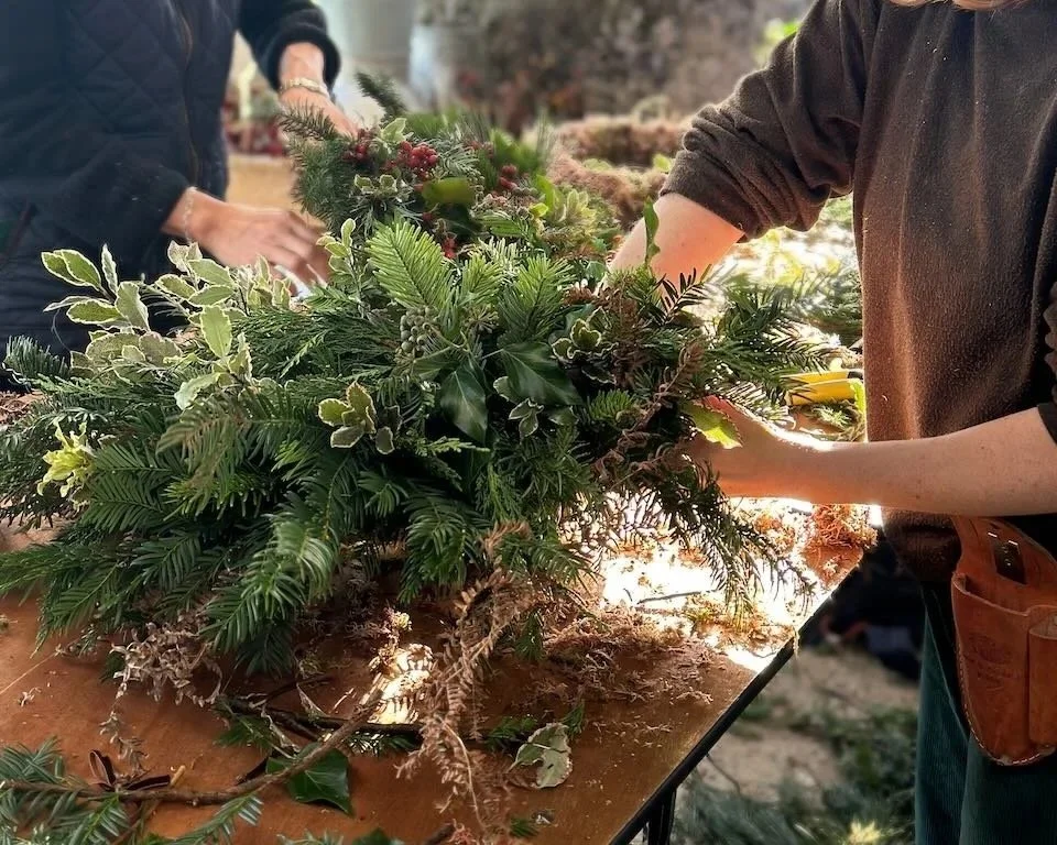 People arranging pine branches, holly, and other greenery for a holiday wreath on a wooden table outside.