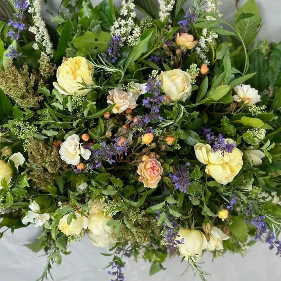 A dense bouquet of mixed flowers including cream-colored roses, small pink roses, and purple blossoms, accented with various green leaves and foliage.