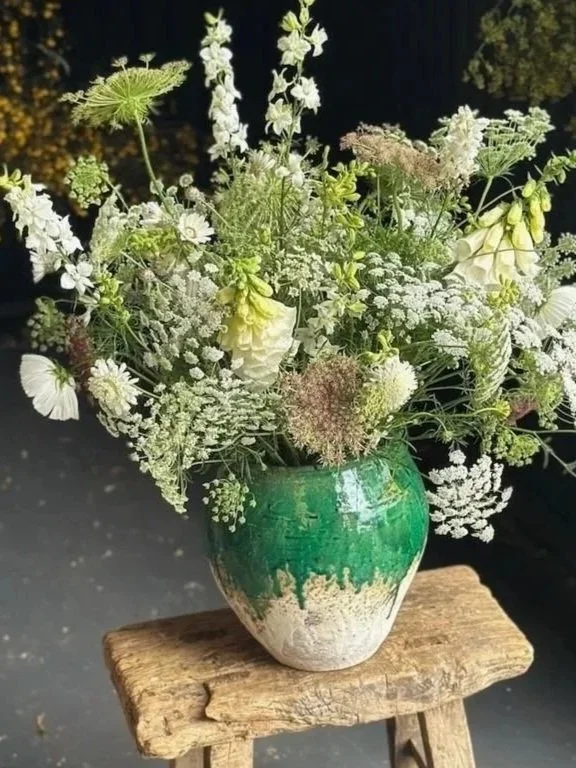 A large bouquet of white and green flowers in a green and cream ceramic vase on a rustic wooden stool.