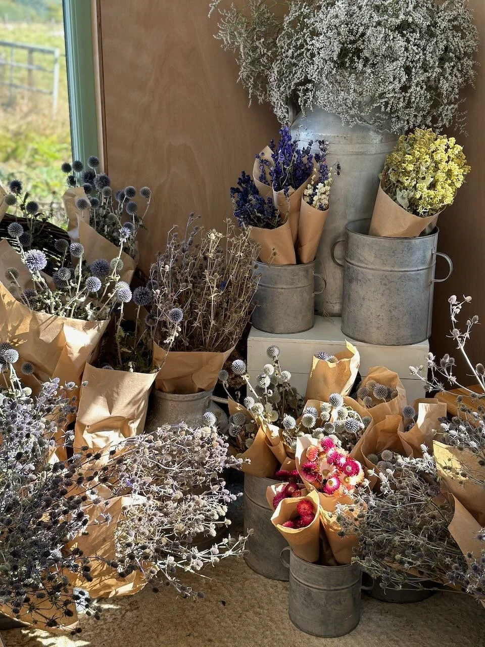 Various bouquets of dried and fresh flowers in paper wrapping, arranged in metal buckets and containers near a window with a green landscape outside.