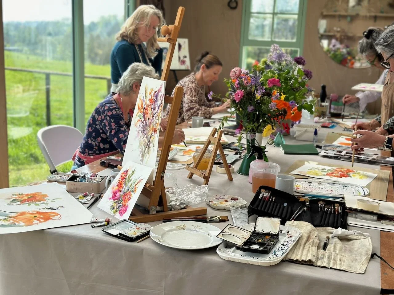 A group of women participating in a watercolor painting class around a table with floral arrangements and art supplies.