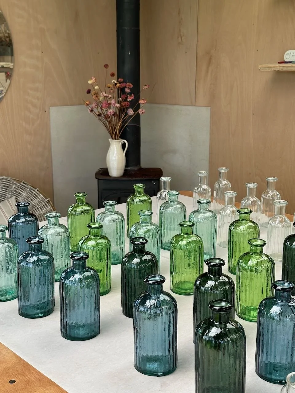 A table filled with various colored glass bottles in shades of blue, green, and clear, with a ceramic vase containing dried flowers in the background.