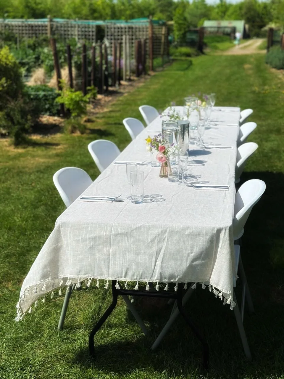 Outdoor garden table set with a white tablecloth, floral centerpieces, glasses, and silverware, situated on a grassy lawn in a backyard with green trees and fencing.