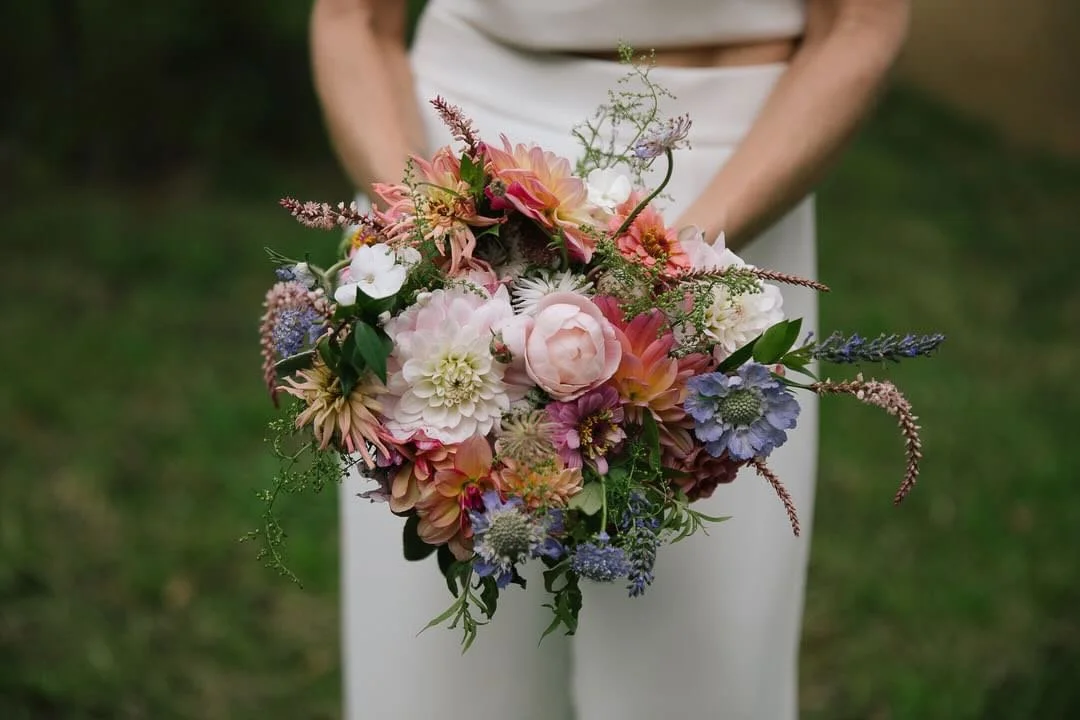 Person holding a colorful bouquet of flowers, including dahlias, roses, and other mixed blooms, with a blurred green outdoor background.