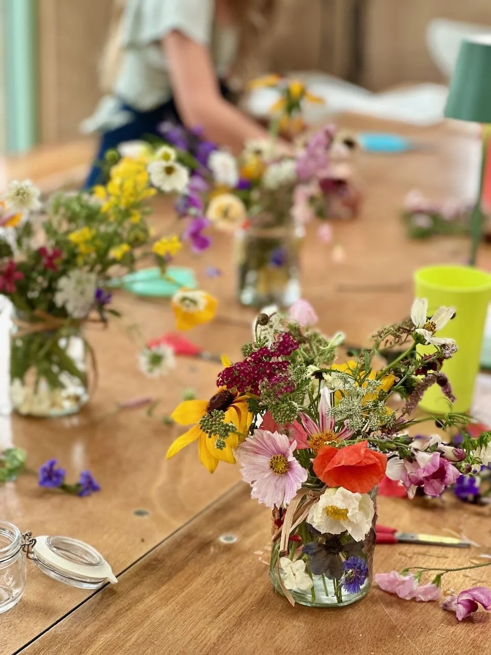 Colorful mixed flower bouquet in a jar on a wooden table with various flowers, scissors, and craft supplies around, with a person working in the background.