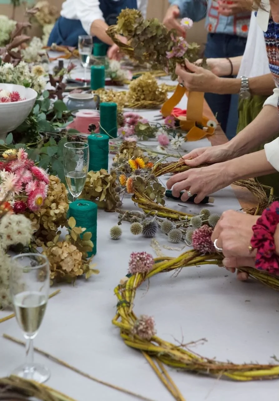 People participating in a wreath-making workshop with floral arrangements, greenery, and candles on the table.