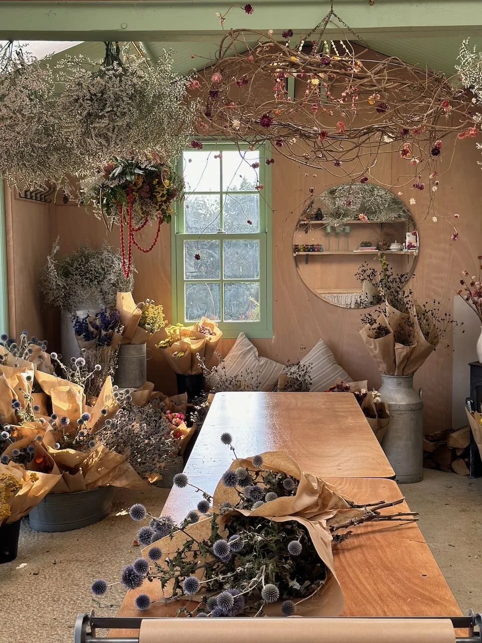 A cozy interior filled with various flower arrangements and dried plants, with a rustic wooden table in the center, a window with green trim letting in natural light, and a round mirror on the wall reflecting shelves and decorations.