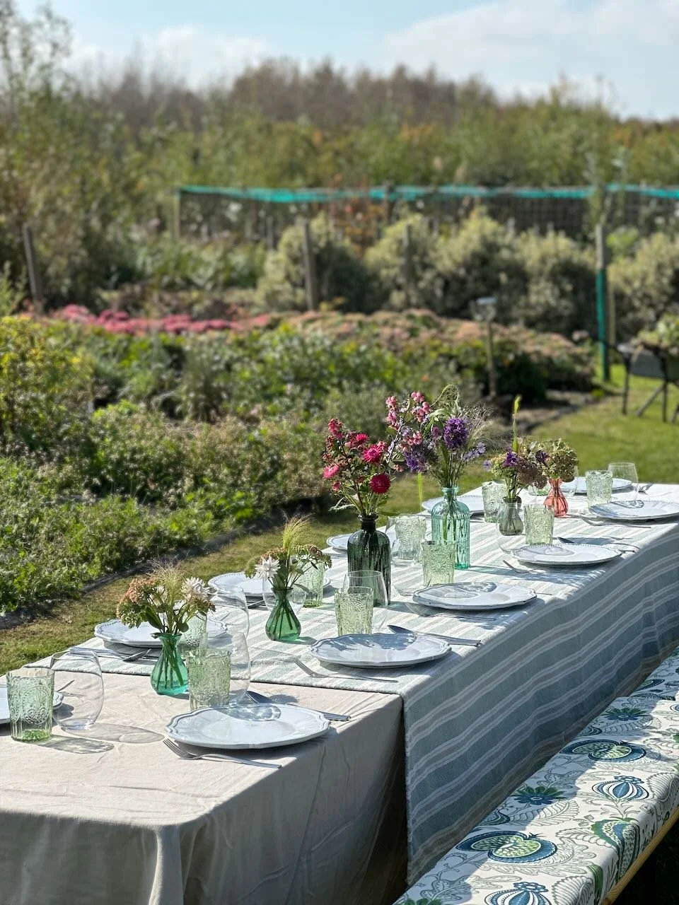 Outdoor dining table set with plates, glasses, and colorful flower vases, surrounded by greenery and flowering bushes.