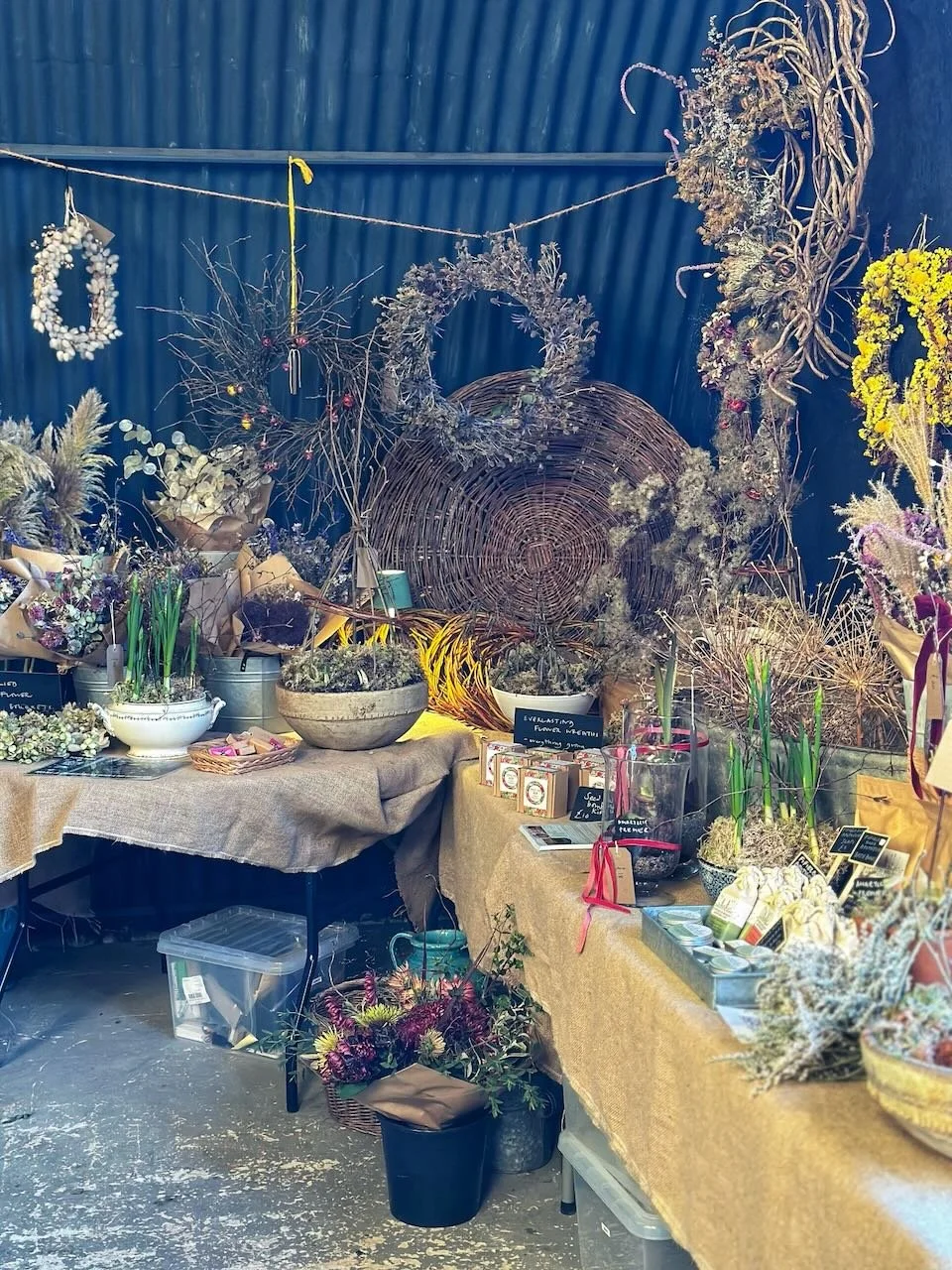 Display of dried flower wreaths and arrangements on tables at a floral shop or market, with wicker wreath frames and various floral supplies.