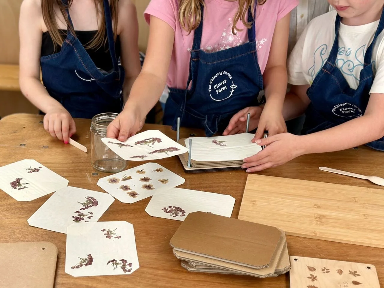 Children participating in a botanical activity, examining pressed flowers and leaves, and placing them on paper sheets on a wooden table, wearing aprons from The Clipping Narthex Flower Farm.