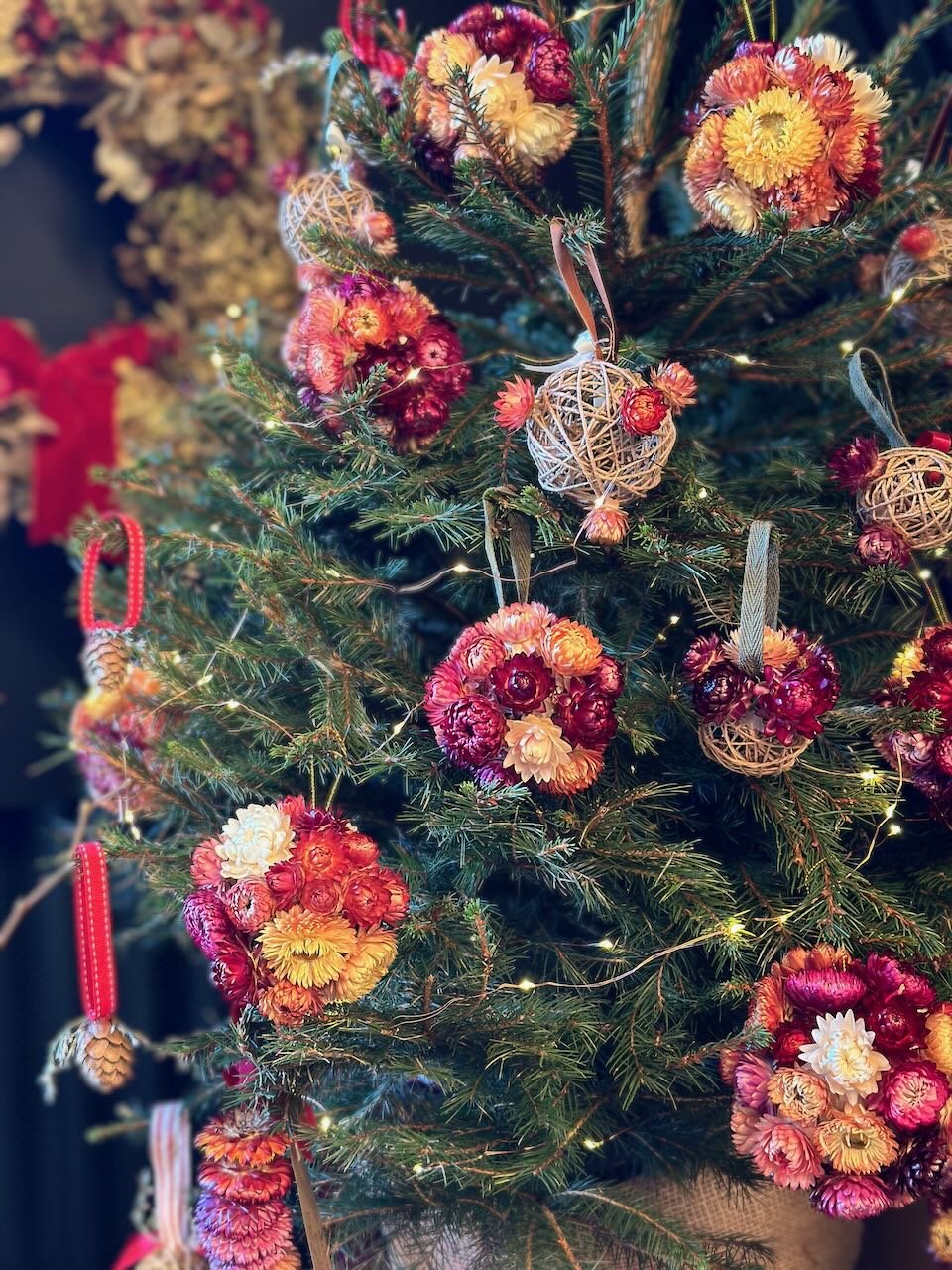 A decorated Christmas tree with string lights, floral ornaments, and woven ball ornaments, featuring red, peach, yellow, and cream-colored flowers and berry accents.