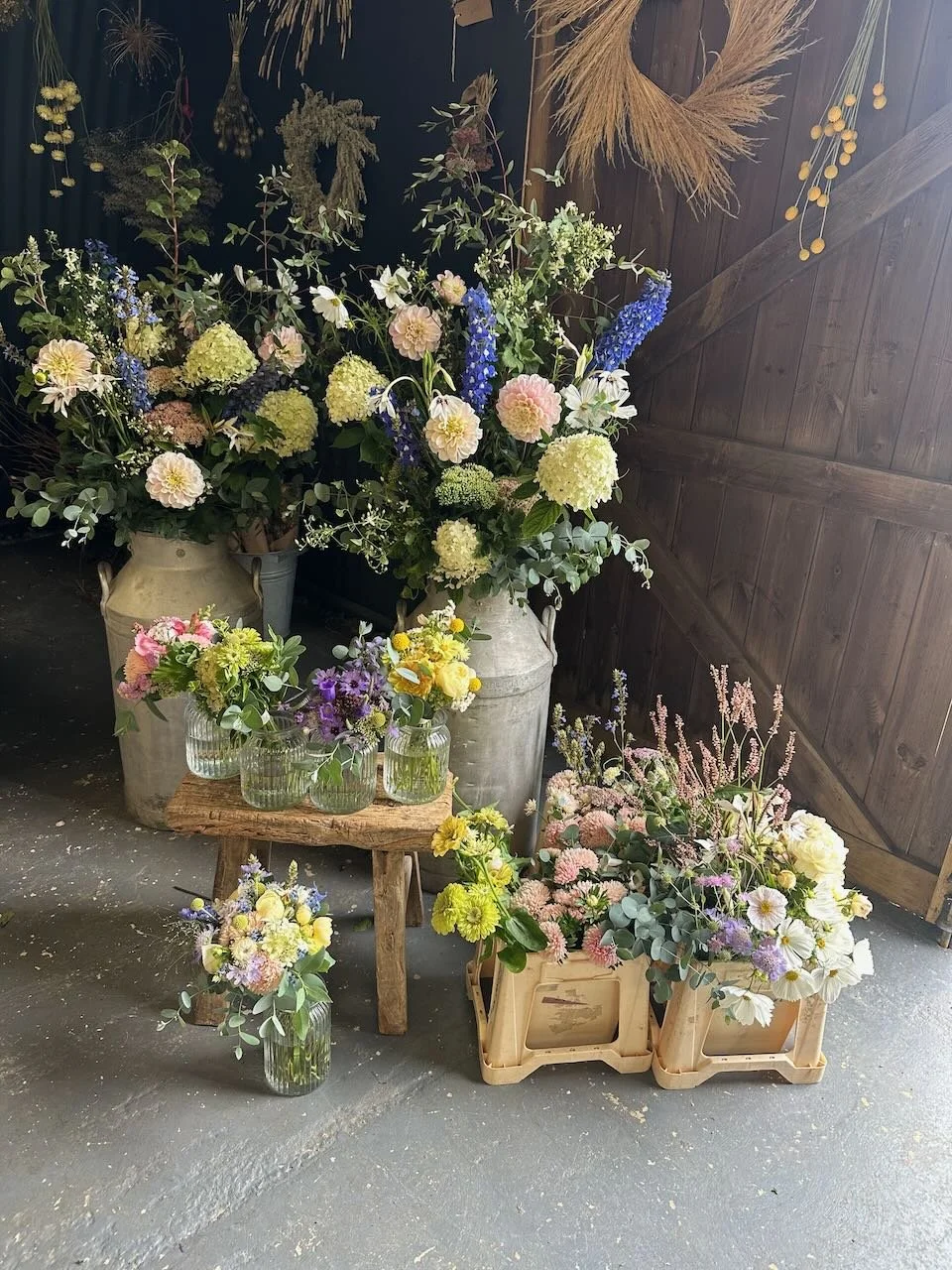 Arrangement of various fresh flowers in vases and crates on rustic wooden furniture against a dark wooden wall.