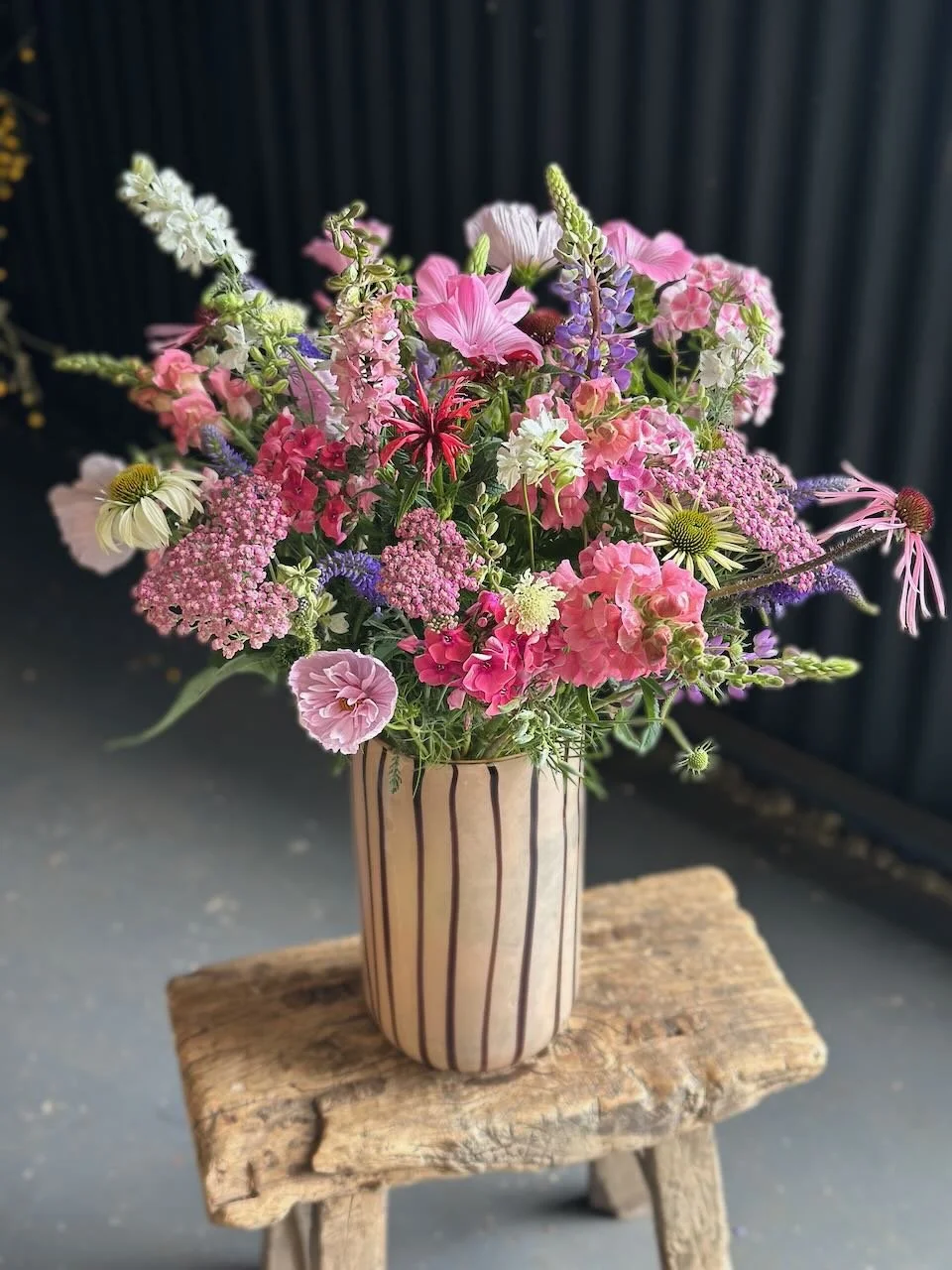 A colorful bouquet of various pink, purple, white, and green flowers in a tall striped vase on a rustic wooden stool.