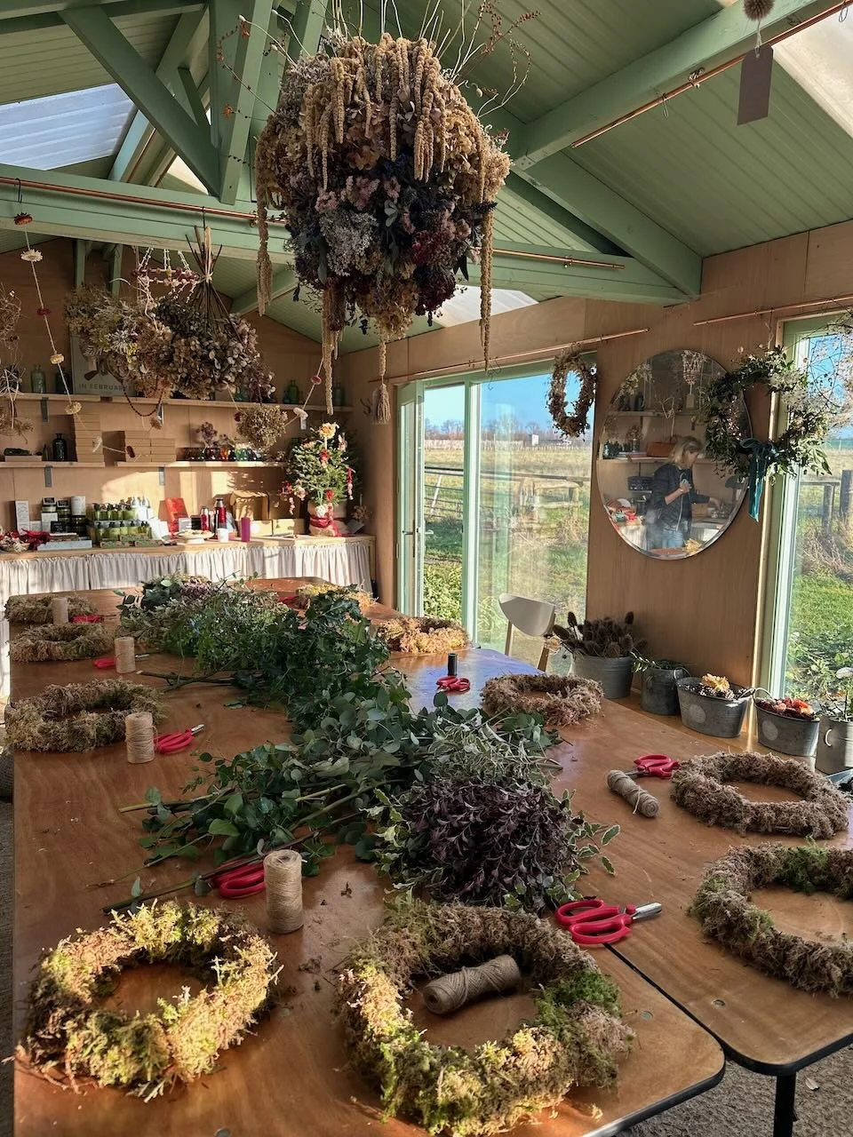 A workshop room decorated with dried flowers and moss wreaths in progress. The table is covered with natural greenery, scissors, and twine. There are shelves with supplies, large windows showing an outdoor field, and hanging dried floral arrangements
