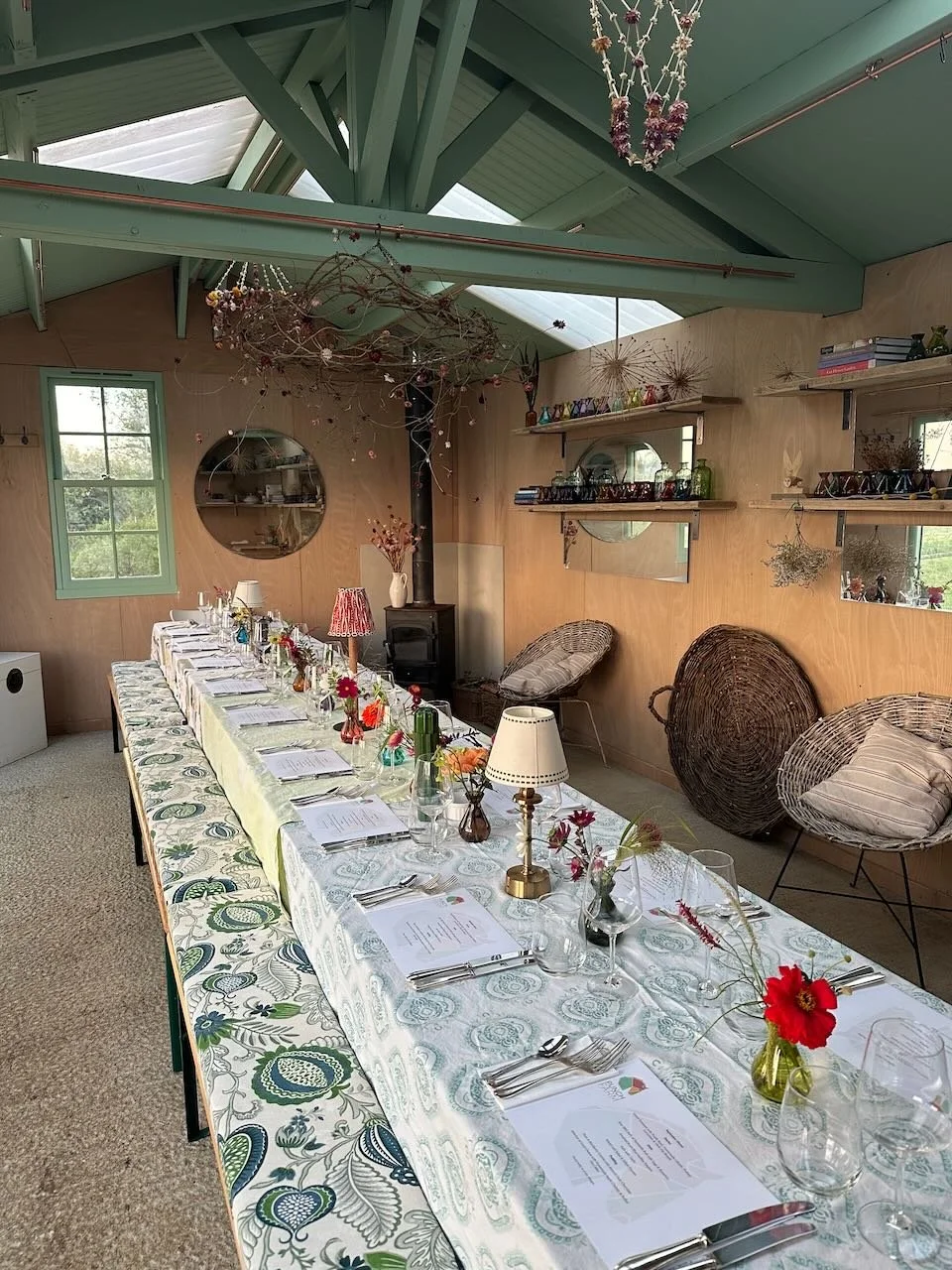 Long dinner table set with plates, glasses, utensils, and small vases with flowers in a rustic room with wooden walls, shelves, and decorative branches hanging from the ceiling.