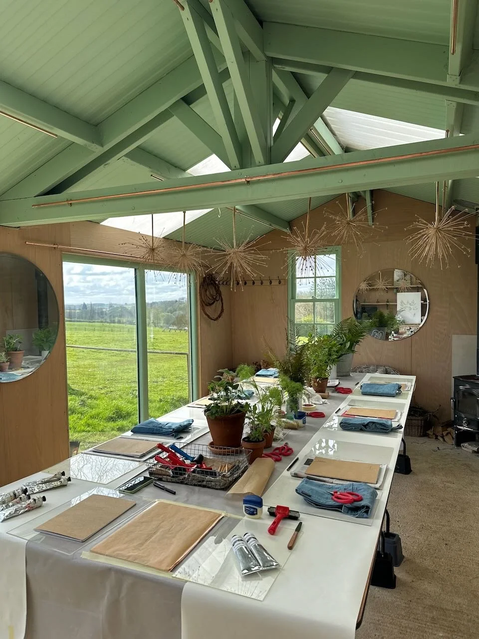 Interior of a craft or workshop space with a long white work table, potted plants, scissors, tools, and blue jeans laid out on the table. Light green painted wooden beams and ceiling, with windows showing a green field outside.